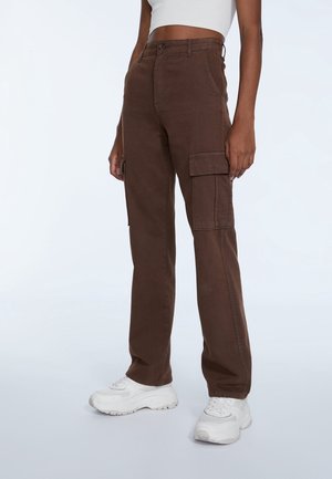 Pantalons cargo marron avec une coupe slim, dotés de poches latérales, d'une fermeture éclair et d'un tissu en mélange de coton. Portés avec des chaussures de sport blanches.