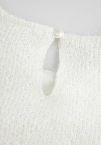 Biały sweter o fakturze z okrągłym otworem przy szyi, zapinany na mały biały perłowy detal. Miękki, puszysty materiał z włóczki.