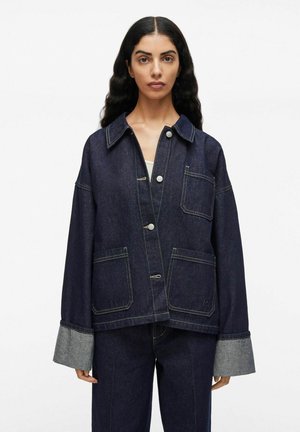 Frau trägt eine dunkelblaue Jeansjacke mit drei vorderen Knöpfen, drei Taschen und breiten umgeschlagenen Ärmeln, kombiniert mit passenden Jeans.