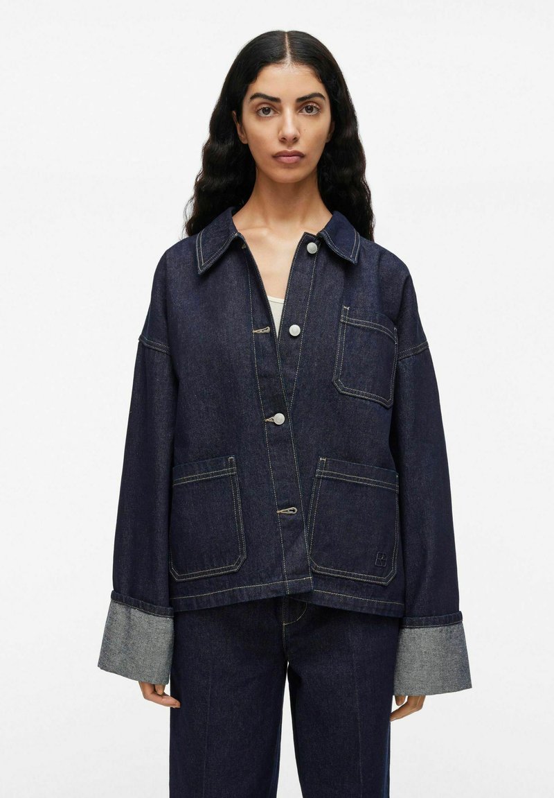 Vrouw in een donkerblauwe denim jas met drie knopen aan de voorkant, drie zakken en brede omgeslagen manchetten, gecombineerd met passende denim broek.