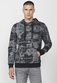 Hoodie u crno-sivoj uzorku s kapuljačom na vezanje, prednjim džepom i rebrastim manžetama. Sadrži geometrijski dizajn s različitim teksturama.