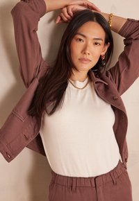 Giacca marrone sopra una camicia bianca, con scollo rotondo. Il modello indossa una collana d'oro e ha lunghi capelli neri e lisci.