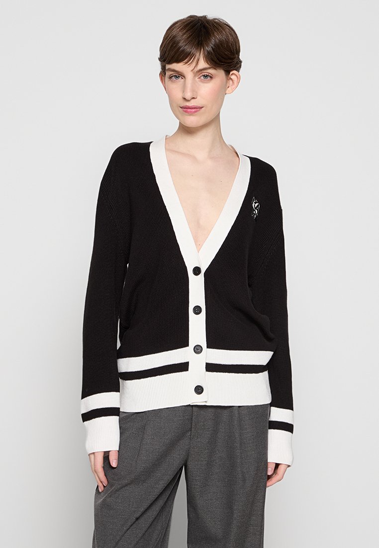 Cardigan noir et blanc avec un décolleté en V profond, cinq boutons, poignets et ourlet rayés, texture côtelée et un petit accent décoratif.