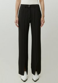 Zwarte wijde broek met vooropeningen, gemaakt van een gladde stof, met een knoopsluiting en twee voorkralen. Draagt met witte laarzen.