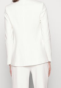 Biały dopasowany blezer o dopasowanym tylnym kroju, z subtelnymi detalami szwów i gładką teksturą. Posiada długie rękawy oraz zarysowaną sylwetkę.