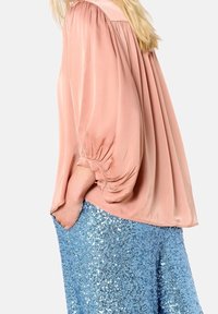 Woman wearing loose, long-sleeve pink blouse with gathered cuffs and sparkly blue sequin pants, hand in pocket, against plain background.