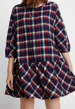 Vrouw draagt een navy, rood en wit geruite jurk met driekwart mouwen en een uitlopende rok, gecombineerd met een ronde gouden hangersketting.