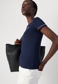 T-shirt ajusté bleu marine avec des bordures rouges et blanches, accompagné d'un sac fourre-tout en cuir noir. Pantalons blancs avec des poches visibles.