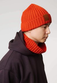 Pomarańczowa dziergana czapka z prążkowanym wzorem i skórzaną naszywką z logo, zestawiona z pasującym prążkowanym pomarańczowym szalikiem, noszona na ciemnej bluzie z kapturem.