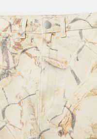Pantalon beige avec motif abstrait de taches de peinture marron et grises, doté de passants de ceinture, d'une fermeture par bouton argenté et de détails de couture visibles.