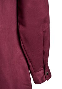 Pikend käised sügavast burgundia värvi riidest, millel on nööpidega mansett ja pehme, nahataoline tekstuur valgel taustal.