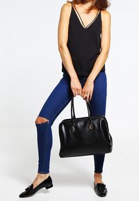 Sac à main en cuir noir avec des poignées doubles, fermeture éclair et détails en quincaillerie décoratifs. Modèle portant un haut noir et un jean skinny bleu.