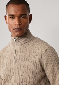 Beige Pullover mit Reißverschluss und strukturiertem Zopfmuster, ausgestattet mit einem gerippten Kragen und Bündchen, aus weichem Material gefertigt.