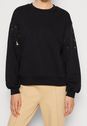 Vrouw draagt een zwart top met lange mouwen en kantuitgesneden details op de mouwen, gecombineerd met beige broek, staand tegen een effen achtergrond.