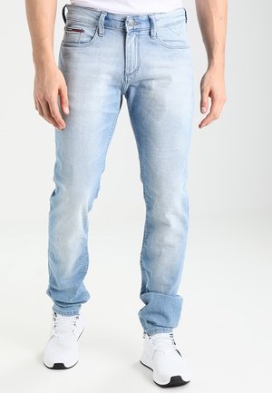 Homme portant un jean slim bleu clair délavé avec des revers roulés et des baskets blanches, debout devant un fond uni.