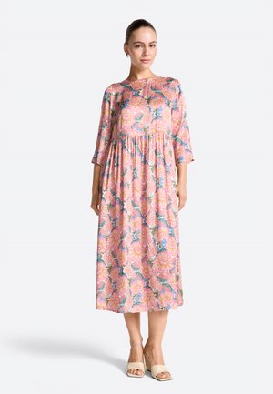 Frau trägt ein midilanges pinkes Kleid mit großem Blumenmuster, 3/4-Ärmeln, kombiniert mit beigen Sandalen mit offenem Zehenbereich und Blockabsatz.