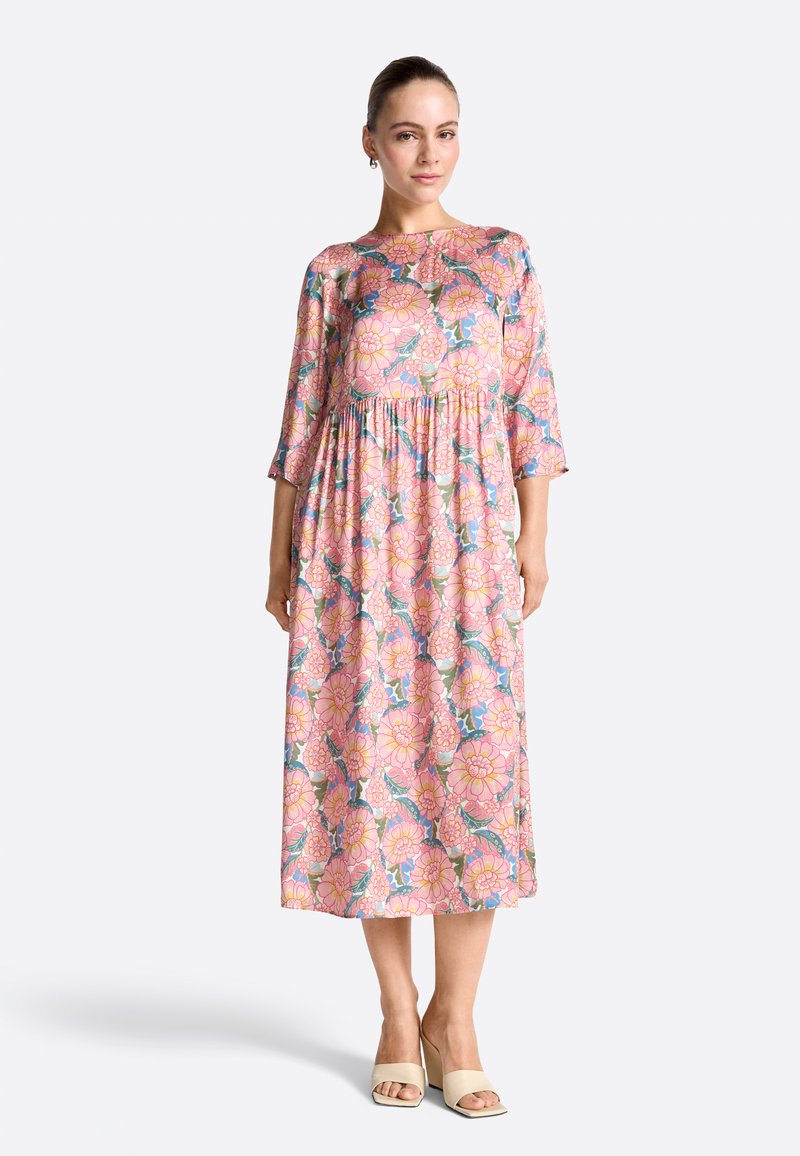 Femme portant une robe midi rose à gros motifs floraux, manches 3/4, associée à des sandales à talons blocs ouvertes beige.