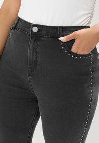 Svarta denimjeans med hög midja, som har en metallknapp, sido fickor och silvernitar längs fickkanternas kant för extra detalj.