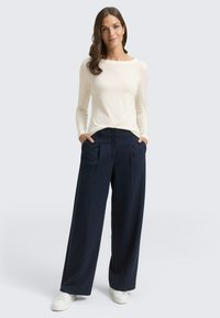 Lichtcrème langemouwen top gecombineerd met marineblauwe wijde broek. De broek heeft plooien aan de voorkant, het model staat met de handen in de zakken. Witte schoenen.