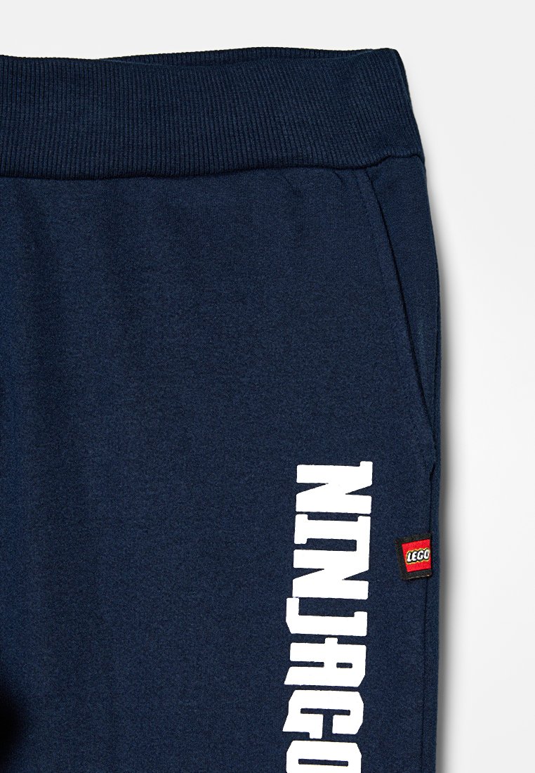 Pantalons de survêtement bleu marine avec taille côtelée, grand texte blanc "NINJAGO" sur un côté, et une petite étiquette du logo LEGO rouge. Texture de tissu douce.