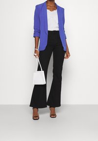 Žena v modrém blejzru, bílém topu a černých rozšířených kalhotách, držící malou bílou kabelku a oblečená v černých podpatcích. Minimalistické pozadí.