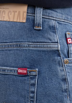 Close-up van de achterzak van een blauwe spijkerbroek met een rood label van het merk "BIG STAR" en zichtbare lussen voor de riem en een leren merkembleem op de tailleband.
