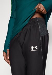 Μαύρο αθλητικό παντελόνι από ελαφρύ ύφασμα με ελαστική μέση, με λευκό λογότυπο Under Armour και υφασμάτινη πλαϊνή λεπτομέρεια.