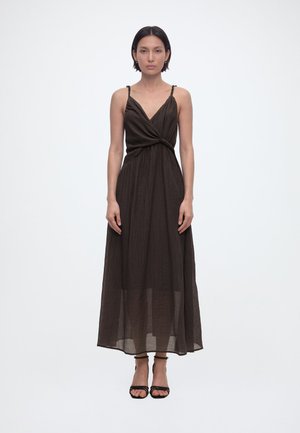 Frau steht frontal, trägt ein ärmelloses schwarzes Wickel-Midikleid und schwarze Riemchenabsätze vor weißem Hintergrund.