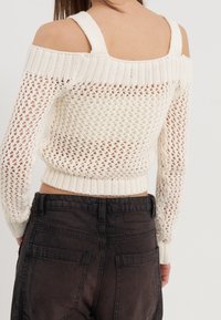 Pull blanc en maille avec épaules dénudées, présentant un motif texturé et un ourlet côtelé. Associé à un pantalon foncé taille haute.