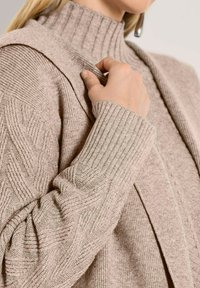 Frau trägt einen beigen gerippten Rollkragenpullover und eine passende strukturierte Strickjacke, hält die Jacke mit einer Hand am Kragen.