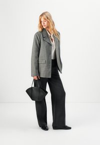 Blazer grigio in misto lana con revers ampi, abbinato a pantaloni neri a vita alta; il modello tiene in mano una borsa strutturata nera.