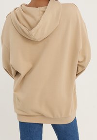 Béžová oversized mikina s kapucí na stažení a bočními švy. Měkká textura látky; má žebrované manžety a lem, stylizována s modrým denimem.