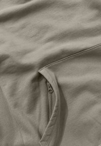 Beige sweatshirtstof met een zachte textuur, voorzien van een ritszak op de zij voor extra functionaliteit en detail.