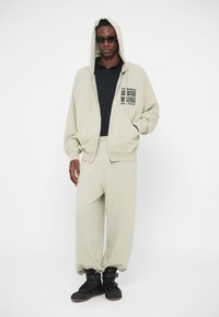 Béžová mikina s predným vreckom a logom, skombinovaná s béžovými nohavicami. Čierne tričko pod ňou a čierna obuv so zapínaním.