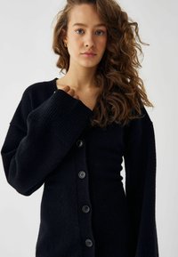 Melnā džemperis ar V veida kakla izgriezumu, ribotām platām piedurknēm un sešām pogām. Mīksta tekstūra ar pielāgotu piegriezumu un brīvu siluetu.