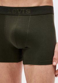 Κοντινό πλάνο ατόμου που φοράει σκούρα πράσινα μπόξερ Levi's, με εμφανές το λογότυπο στη μέση και το χέρι να ακουμπάει στο μηρό.