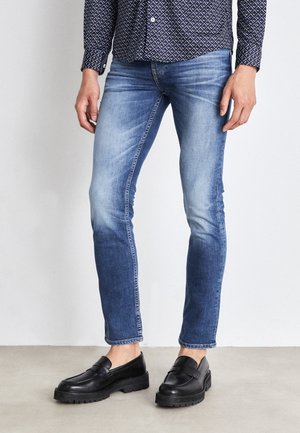 Homme portant un jean skinny délavé bleu clair et bleu foncé avec des mocassins en cuir noir et une chemise à manches longues à motifs.