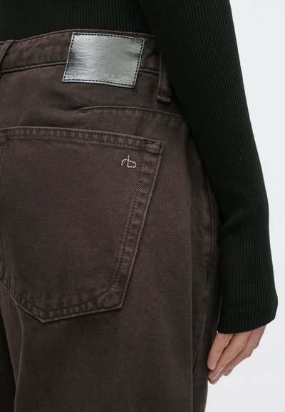 Jeans de ganga castanha com um bolso traseiro e um emblema de logótipo em couro preto. O tecido tem um ligeiro brilho, com detalhes sutis de costura.