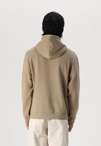 Beige pullover hoodie with a drawstring hood, soft texture, ribbed hem and cuffs, and a relaxed fit, worn with light-colored pants.
