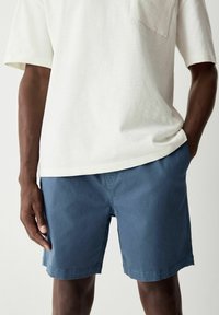 Camiseta de manga corta gris claro con un bolsillo, combinada con pantalones cortos azul marino. Los pantalones cortos tienen una textura suave y un corte recto.