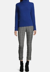 Kvinna iklädd en blå polotröja, grå rutiga slim-fit byxor och svarta ankelstövlar som står framför en vit bakgrund.