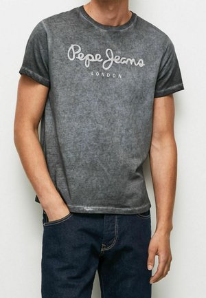 Mężczyzna ubrany w wyprany szary T-shirt Pepe Jeans London i ciemnoniebieskie dżinsy, z jedną ręką w kieszeni, stojący na jednolitym białym tle.