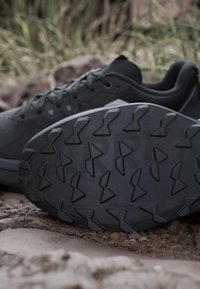 Scarpa da trail running nera con suola strutturata e motivi angolari antiscivolo su terreno roccioso all'aperto.