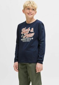 T-shirt a maniche lunghe di colore navy con stampa grafica rossa e bianca. Realizzato in morbido cotone; design con collo rotondo. Indossato con pantaloni verde oliva.