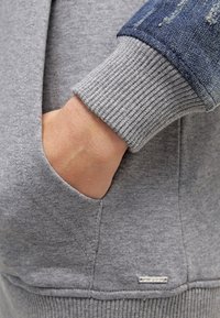 Felpa grigia con polsini a coste e tasca frontale. Presenta dettagli in denim sulle maniche e un piccolo logo metallico vicino all'orlo.