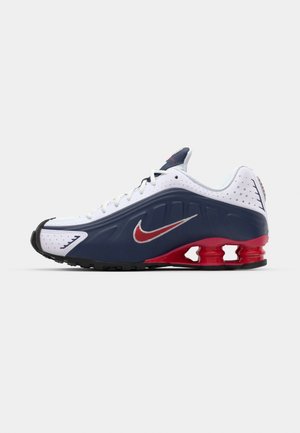 Scarpa sportiva bianca e blu navy con accenti rossi, pannelli in rete, collo imbottito e design distintivo della suola con unità di imbottitura visibili.