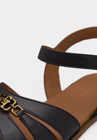 Μαύρο δερμάτινο σανδάλι με χρυσή διακοσμητική αλυσίδα στο μπροστινό λουρί και καφετί μαξιλαράκι στο πέλμα, εμφανίζεται σε στενή λήψη.