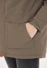 Brun, texturerad jacka med hög krage, framfickor och kardborreackcenter vid ärmarna. Utformad för värme och funktionalitet.