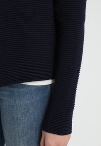 Námořnický pletený svetr s dlouhými rukávy, strukturovaná látka a lemování nad pasem, spárovaný se světle modrými džíny.