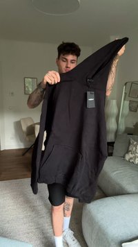 Giovane uomo con tatuaggi tiene in alto una felpa nera con tasca frontale e etichetta all'interno di un soggiorno con divano e poltrona.